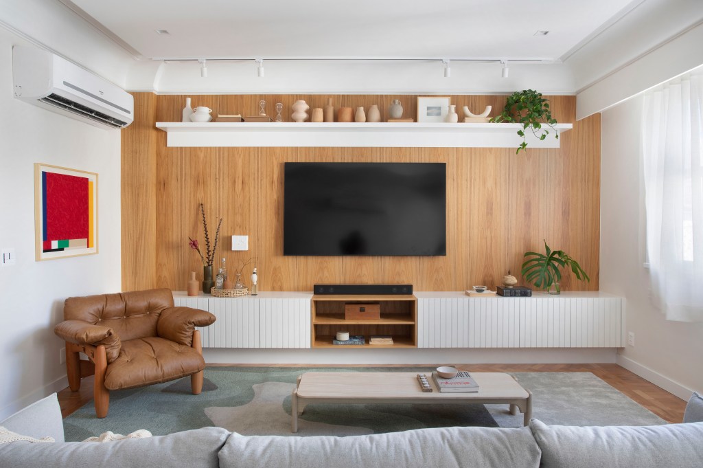 Projeto de Henrique Ramalho. Na foto, sala de estar com piso de taco, piano, sofá cinza azulado e parede revestida de madeira.