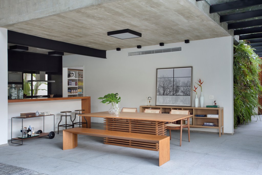 A varanda desta residência de 511 m² fica dentro de casa. Projeto Sabugosa Arquitetura. Na foto, sala de jantar e cozinha integrada ao jardim com mesa de madeira e despensa.