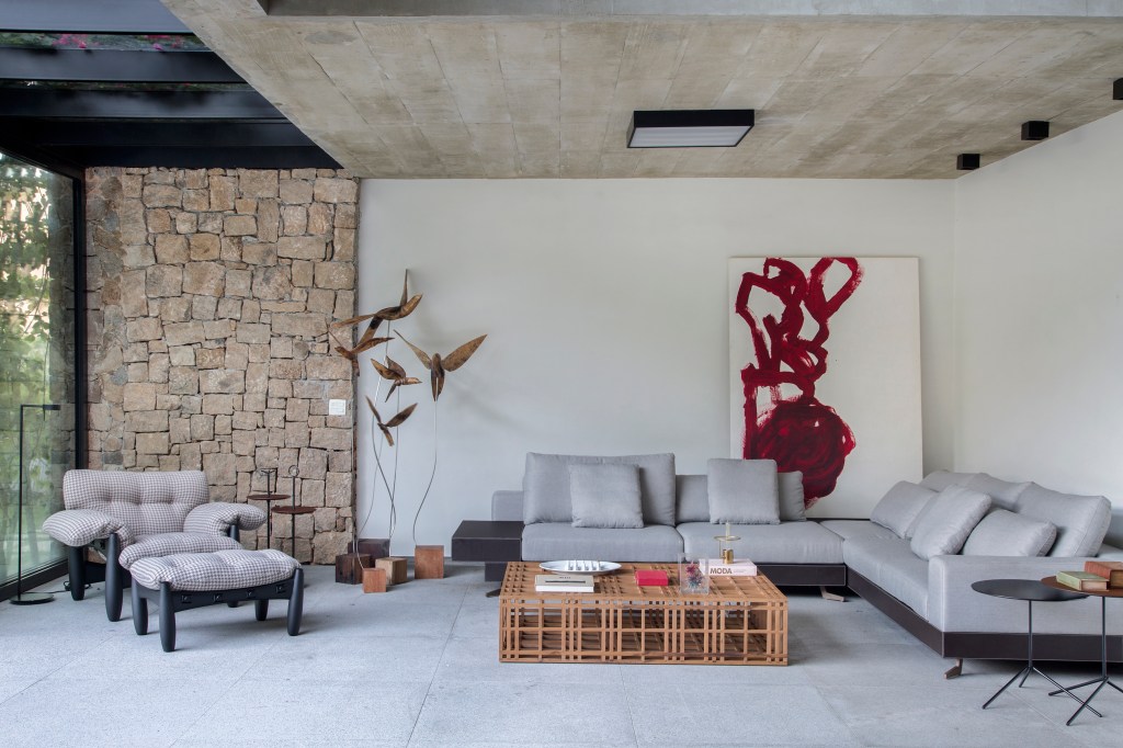 A varanda desta residência de 511 m² fica dentro de casa. Projeto Sabugosa Arquitetura. Sala de estar integrada ao jardim com parede de pedra, poltrona mole e sofá em l.