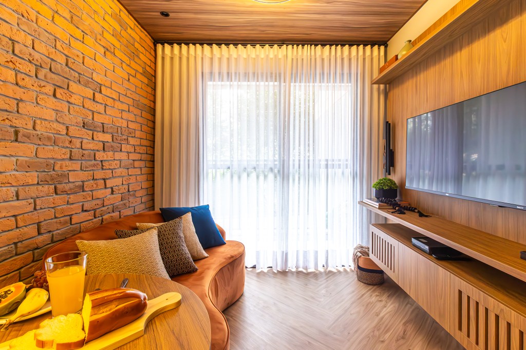 Apê de 39 m² tem cozinha azul, tijolos rústicos e quarto de hóspedes. Projeto de Larice Sena. Na foto, sala de estar com sofá curvo laranja e parede de tijolinho.