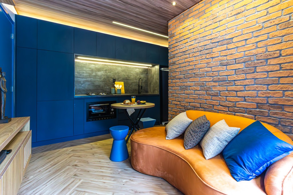 Apê de 39 m² tem cozinha azul, tijolos rústicos e quarto de hóspedes. Projeto de Larice Sena. Na foto, cozinha com marcenaria azul e parede de tijolos rústicos. Sofá curvo laranja.