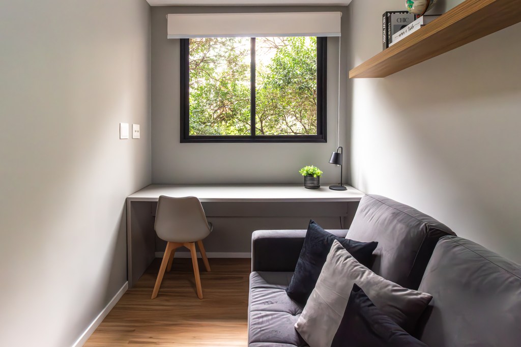 Apê de 39 m² tem cozinha azul, tijolos rústicos e quarto de hóspedes. Projeto de Larice Sena. Na foto, home office com sofá cinza e cortina.