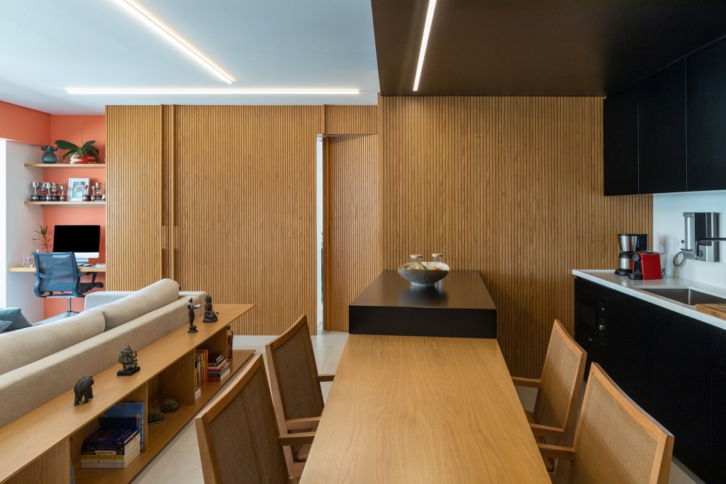 Apê de 90 m² tem cozinha com marcenaria preta e cuba triangular no lavabo. Projeto de Joaquim Meyer. Na foto, sala de estar com jantar integrada, cozinha e painel ripado.