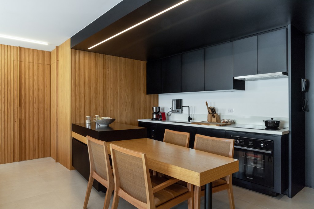 Apê de 90 m² tem cozinha com marcenaria preta e cuba triangular no lavabo. Projeto de Joaquim Meyer. Na foto, sala de estar com jantar integrada, cozinha e painel ripado.