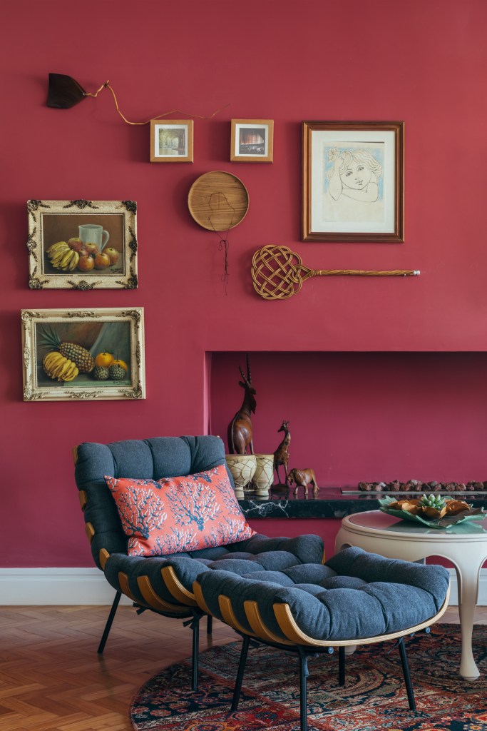 Casa de casal de 70 anos apaixonado por samba inspira-se na brasilidade. Projeto de Ana Cano. Na foto, sala de estar com parede vermelha e poltrona costela com pufe.