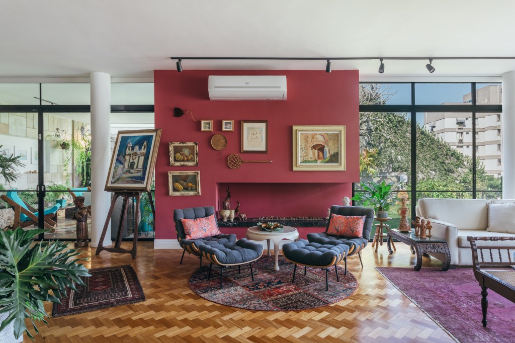 Casa de casal de 70 anos apaixonado por samba inspira-se na brasilidade. Projeto de Ana Cano. Na foto, sala de estar com parede vermelha e poltrona costela com pufe. Piso de taco.