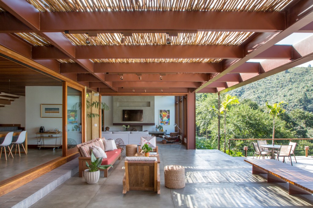 Casa na serra imersa na natureza possui vista espetacular para montanhas. Projeto de Andrea Chicharo. Na foto, varanda com pergolado, tv e lareira.