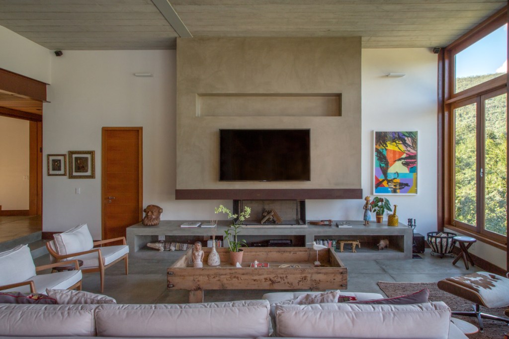 Casa na serra imersa na natureza possui vista espetacular para montanhas. Projeto de Andrea Chicharo. Na foto, sala com vista para a serra, tv, lareira e móveis de concreto.
