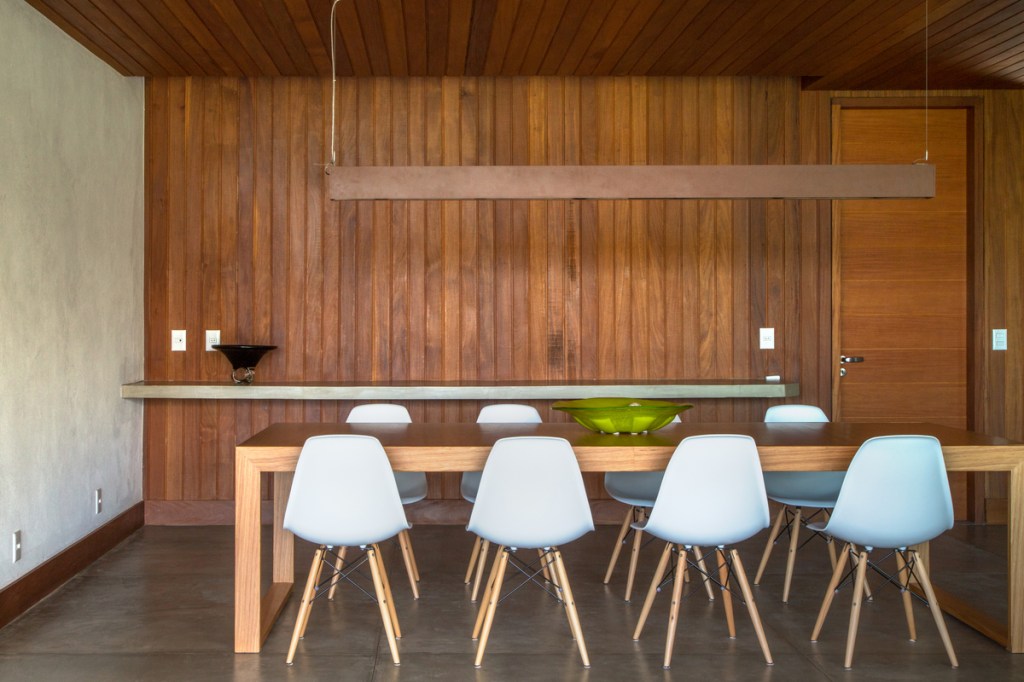 Casa na serra imersa na natureza possui vista espetacular para montanhas. Projeto de Andrea Chicharo. Na foto, sala de jantar com parede de madeira e cadeiras brancas.