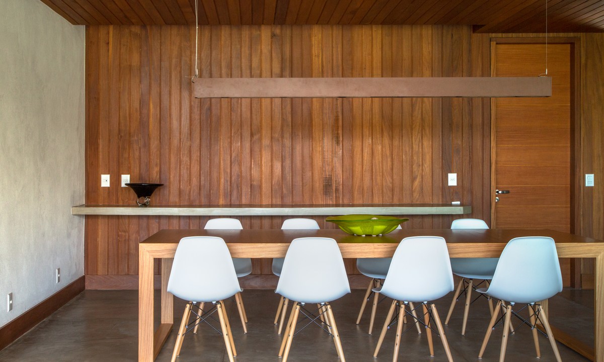 Casa na serra imersa na natureza possui vista espetacular para montanhas. Projeto de Andrea Chicharo. Na foto, sala de jantar com parede de madeira e cadeiras brancas.