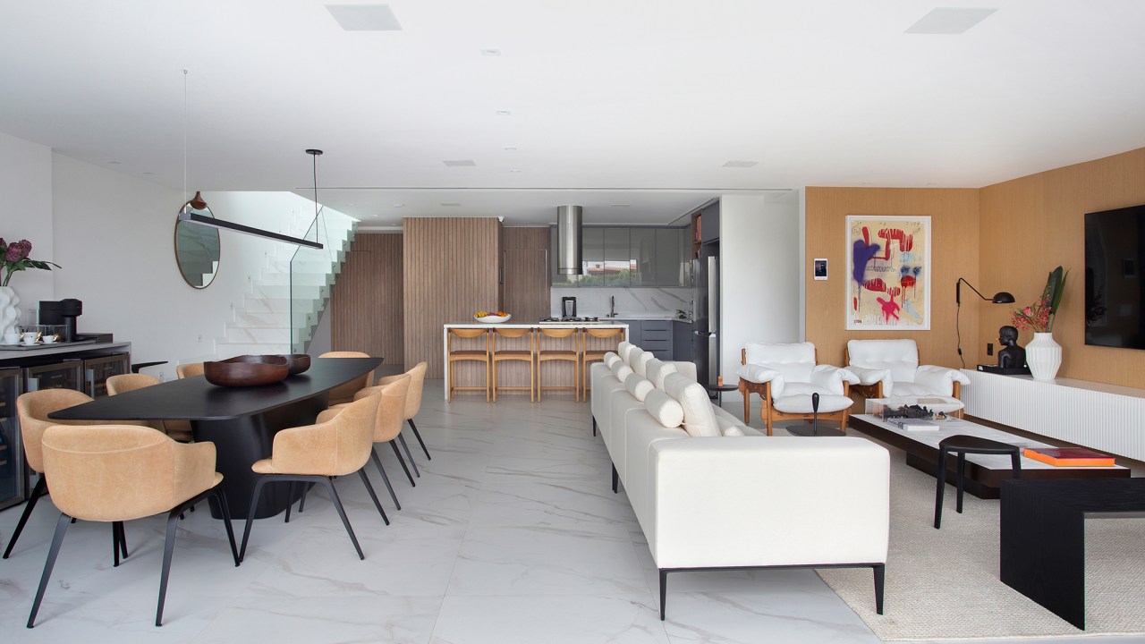 Duplex de 280 m² é minimalista, integrado e repleto de design brasileiro. Projeto de Tom Castro. Na foto, sala de tv com parede de madeira, poltronas e quadro. Sala de jantar com mesa preta e escada branca.