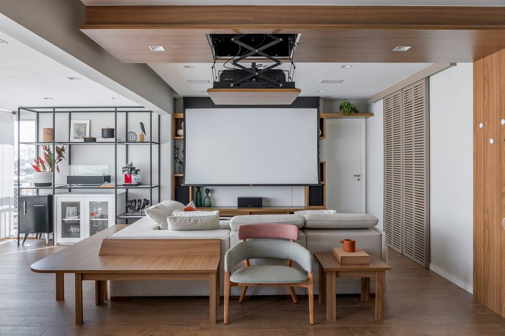 Telão embutido no forro da sala é destaque deste apê de 130 m². Projeto de Ju Matos Arquitetura. Na foto, sala de estar com tv, estante vazada e sofá em L. Telão embutido no forro.