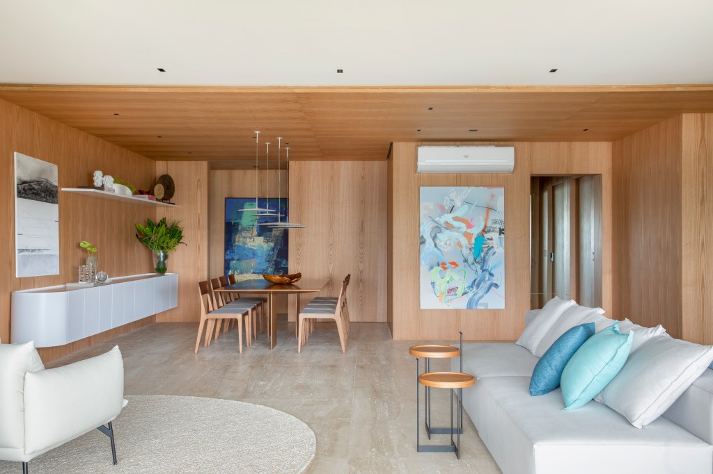 Varanda gourmet espaçosa com ilha travertino natural é destaque em apê. Projeto de Up3 Arquitetura. Na foto, sala integrada com pórtico de madeira e sofá ilha cinza.