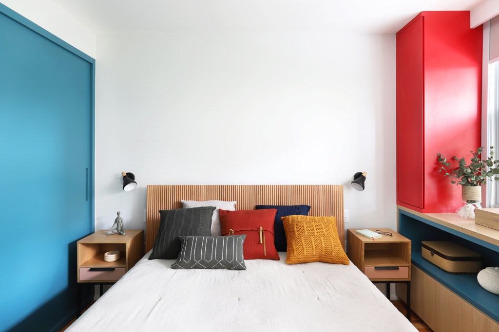 Apê de 90m² tem cozinha rosa, armário vermelho e guarda-roupa azul. Projeto de Studio 92 Arquitetura. Na foto, quarto com cabeceira de madeira, pilar de madeira vermelha.