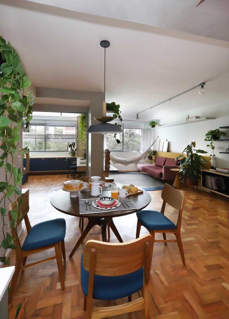 Apê de 90m² tem cozinha rosa, armário vermelho e guarda-roupa azul. Projeto de Studio 92 Arquitetura. Na foto, sala de jantar com mesa redonda, piso de madeira e planta suspensa.