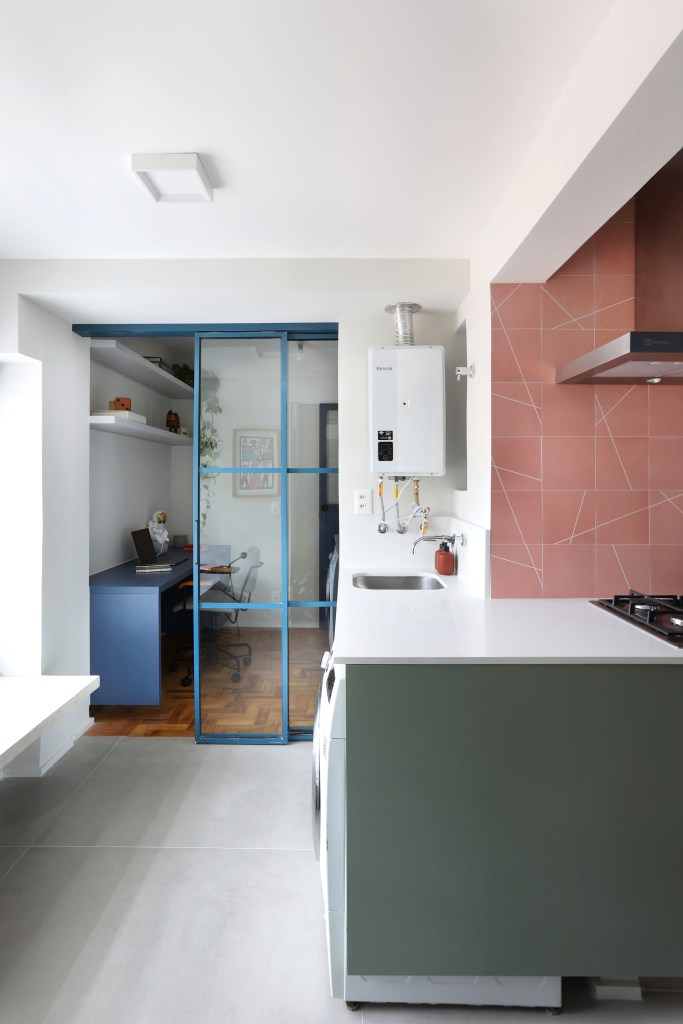 Apê de 90m² tem cozinha rosa, armário vermelho e guarda-roupa azul. Projeto de Studio 92 Arquitetura. Na foto, cozinha integrada com lavanderia e home office ao fundo separado por porta de correr.