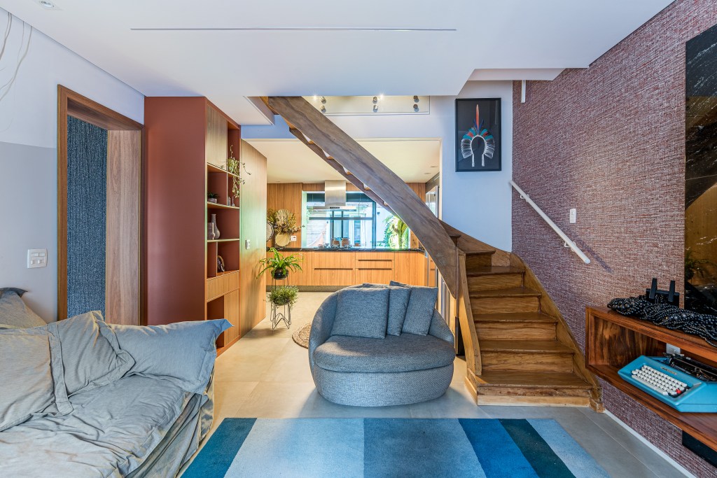 Casa de vila tem jardim com rede, cozinha integrada e muito charme. Projeto de Marcela Fazio Arquitetura. Na foto, sala de estar com sofá cinza, escada e tapete azul.