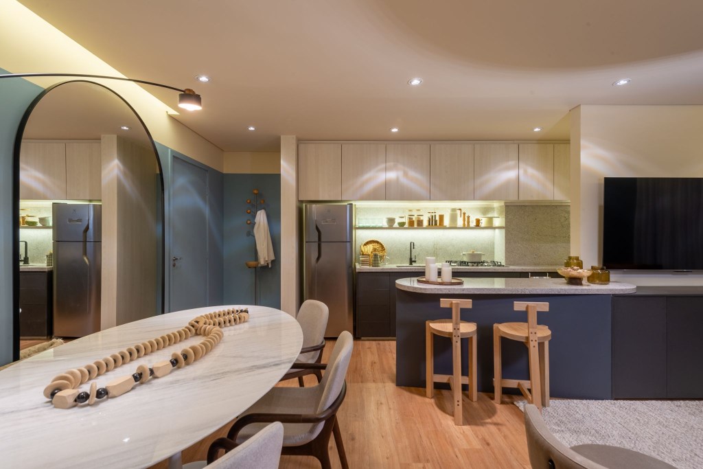 Cores vivas marcam apês de 64 e 85 m² assinados por Todos Arquitetura. Na foto, sala de jantar com espelho, mesa redonda e paredes azuis. Cozinha integrada com balcão.