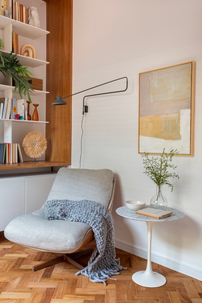 Dicas fáceis para montar um cantinho da leitura em casa. Projeto de Rafael Ramos. Na foto, cantinho de leitura com poltrona cinza, mesinha lateral e luminária de parede.