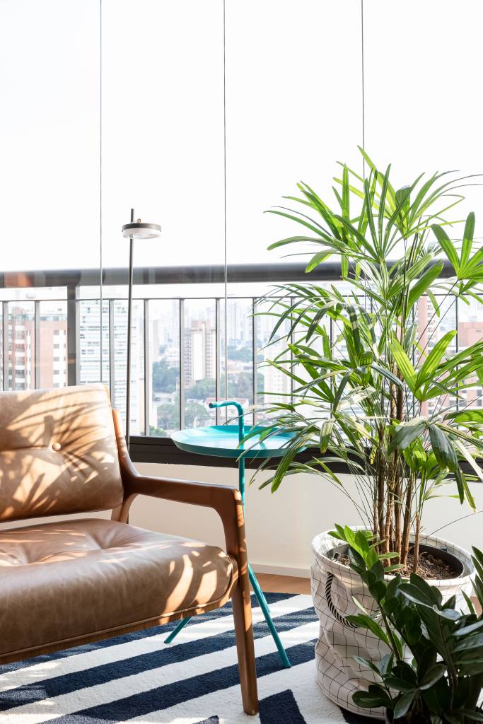 Painel e rack de TV separam living de home office em apê de 75m². Projeto de ISE Arquitetura. Na foto, varanda com tapete azul e branco e poltrona.