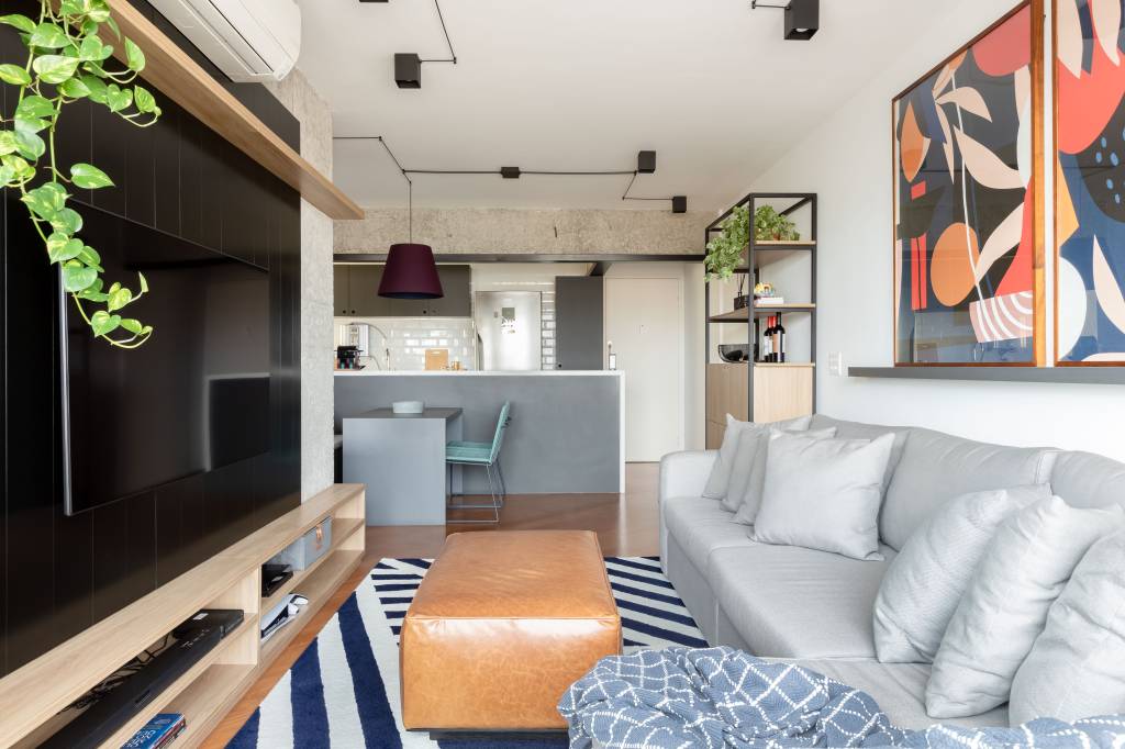 Painel e rack de TV separam living de home office em apê de 75m². Projeto de ISE Arquitetura. Na foto, sala de estar com sofá cinza, tapete azul e branco, pufe e painel de tv preto.