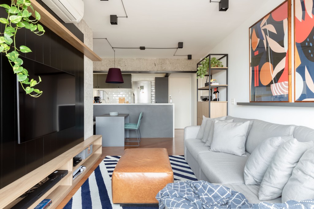 Painel e rack de TV separam living de home office em apê de 75m². Projeto de ISE Arquitetura. Na foto, sala de estar com sofá cinza, tapete azul e branco, pufe e painel de tv preto.