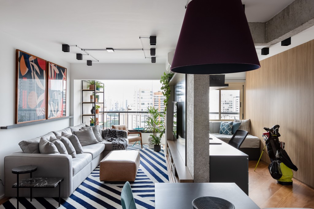 Painel e rack de TV separam living de home office em apê de 75m². Projeto de ISE Arquitetura. Na foto, sala de estar com sofá cinza, tapete azul e branco, pufe e painel de tv preto. Quarto de hóspedes.