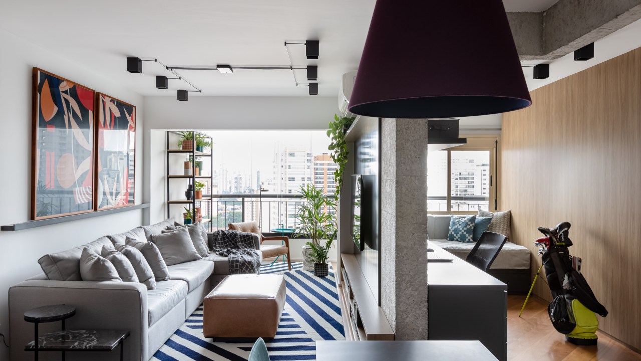 Painel e rack de TV separam living de home office em apê de 75m². Projeto de ISE Arquitetura. Na foto, sala de estar com sofá cinza, tapete azul e branco, pufe e painel de tv preto. Quarto de hóspedes.