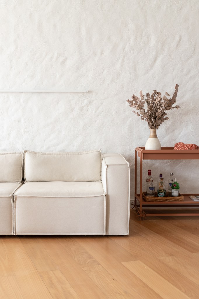 Vigas, reboco aparente e texturas artesanais marcam este apê de 130 m². Projeto de Mariana Monnerat. Na foto, sala de estar com parede texturizada e sofá branco.