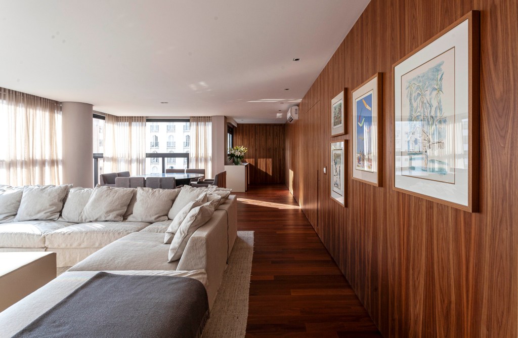 Apê de 174 m² tem decoração clássica e sofisticada com tons neutros. Projeto de David Bastos. Na foto, sala de estar ampla com piso de madeira e parede revestida de madeira com quadros.