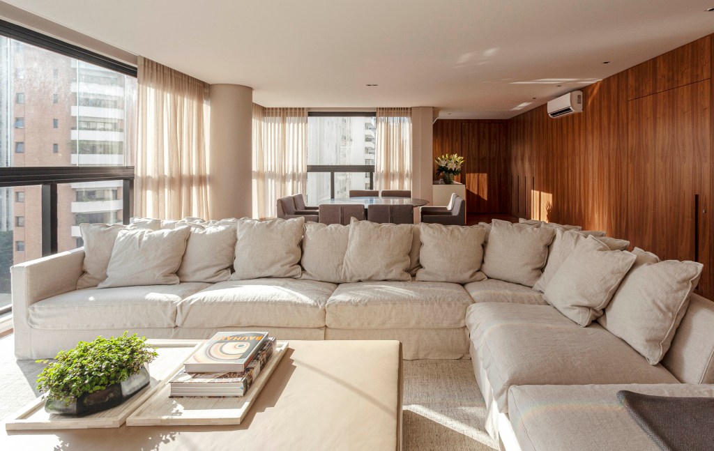Apê de 174 m² tem decoração clássica e sofisticada com tons neutros. Projeto de David Bastos. Na foto, sala de estar com parede revestida de madeira e sofás grandes brancos.