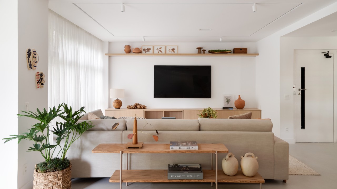Apê de 230 m² ganha décor com tons terrosos e ilha de cozinha com reforma. Projeto de Paula Muller. Na foto, sala de estar em tons neutros, sofá claro, aparador atrás do sofá e planta.