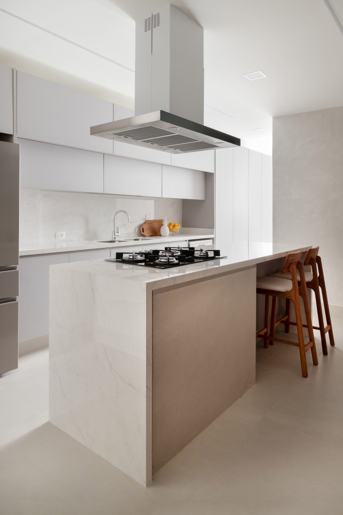 Apê de 230 m² ganha décor com tons terrosos e ilha de cozinha com reforma. Projeto de Paula Muller. Na foto, cozinha minimalista com ilha, banquetas, coifa, marcenaria branca.