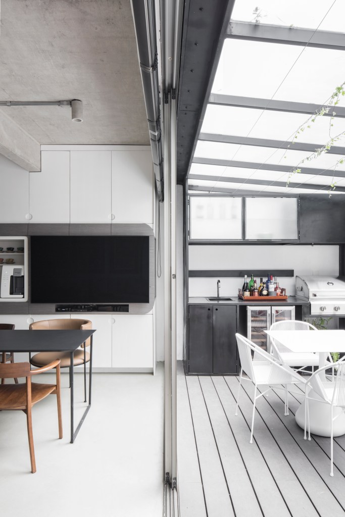 Apê térreo preto e branco tem áreas sociais e de trabalho mescladas. Projeto de Nati Minas & Studio. Na foto, varanda com deck, mesa e cadeiras brancas, cobertura transparente, sofá baixo laranja. Cozinha integrada.