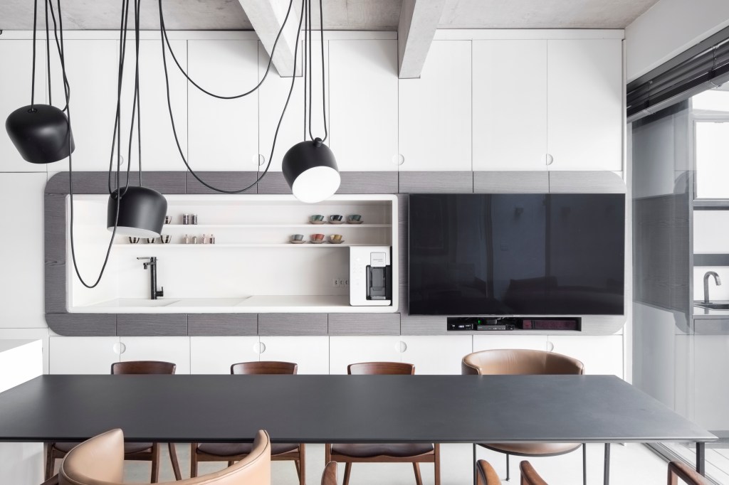 Apê térreo preto e branco tem áreas sociais e de trabalho mescladas. Projeto de Nati Minas & Studio. Na foto, cozinha integrada, luminária preta, mesa preta, marcenaria branca.