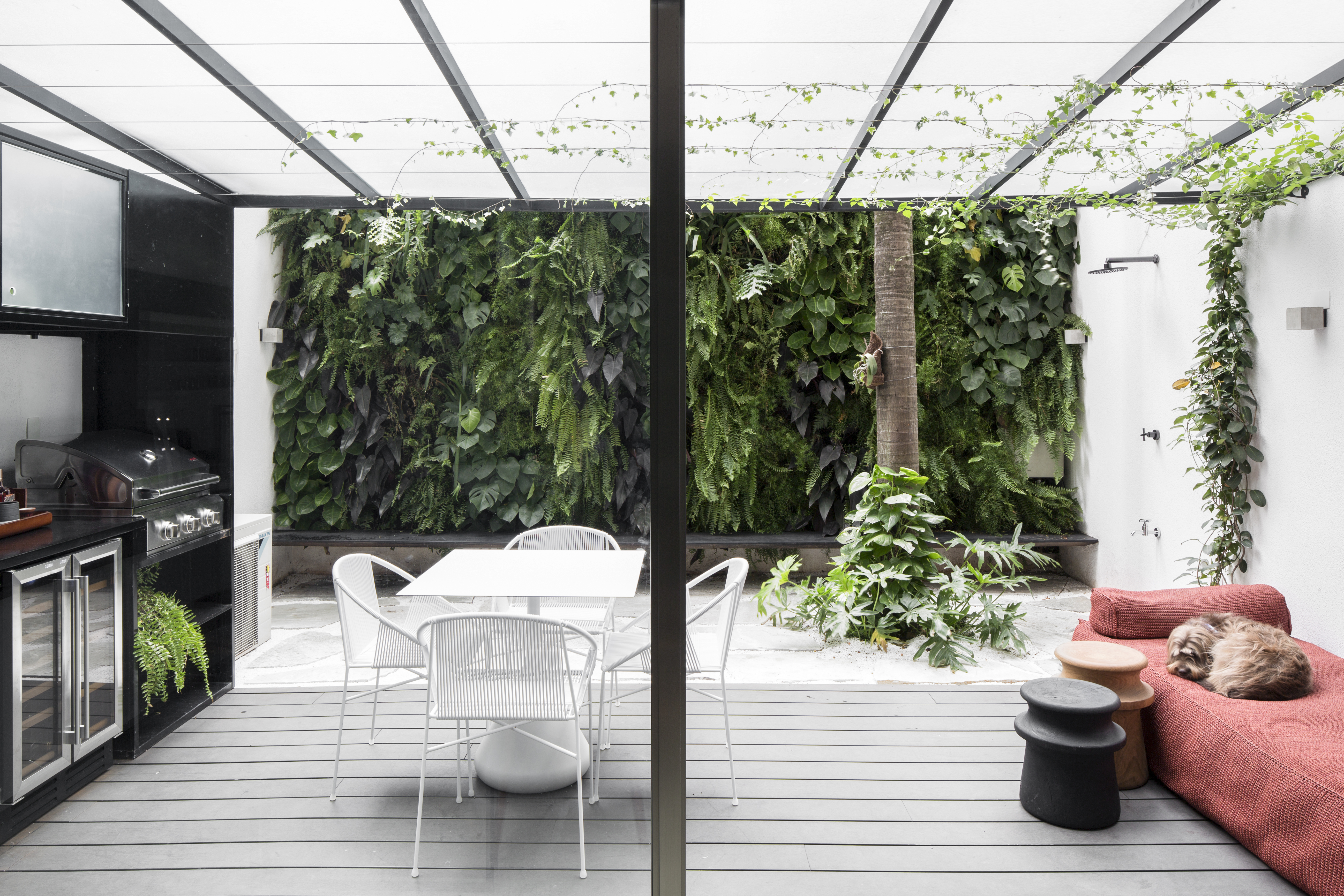 Apê térreo preto e branco tem áreas sociais e de trabalho mescladas. Projeto de Nati Minas & Studio. Na foto, varanda com deck, mesa e cadeiras brancas, cobertura transparente, sofá baixo laranja.