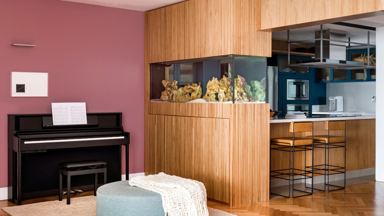 Aquário de peixes funciona como divisória entre sala e cozinha neste apê. Projeto de Vivi Cirello. Na foto, sala, aquário, parede rosa, piano, pufe.