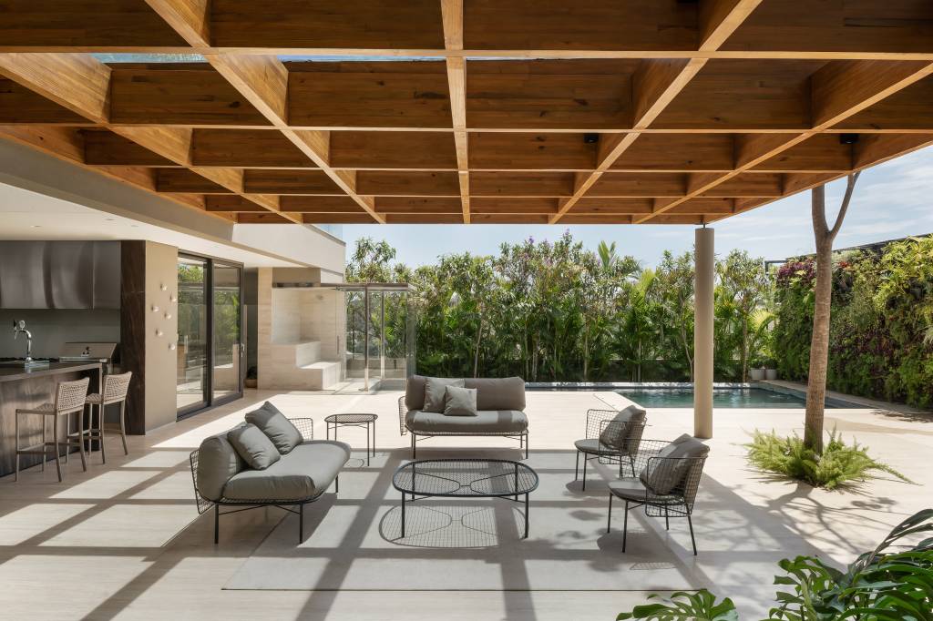 Casa erguida do zero ganha duas cozinhas e muita madeira ripada. Projeto de RF Arquitetura. Na foto, varanda com pergolado de madeira, poltronas cinzas, mesa de centro.