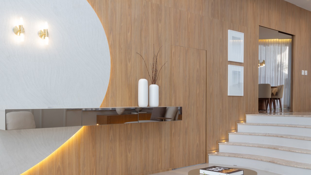 Casa do zagueiro da seleção Gleison Bremer tem estilo minimalista elegante. Projeto de Daniel Santana. Na foto, sala de estar com parede revestida de madeira, espelho e aparador.