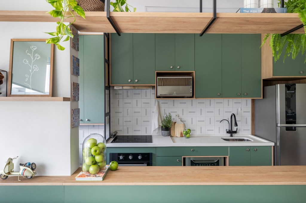 Cozinha verde e granilite no banheiro marca apartamento de 58 m². Projeto de Pixel Arquitetura. Na foto, cozinha americana com marcenaria verde e prateleiras.