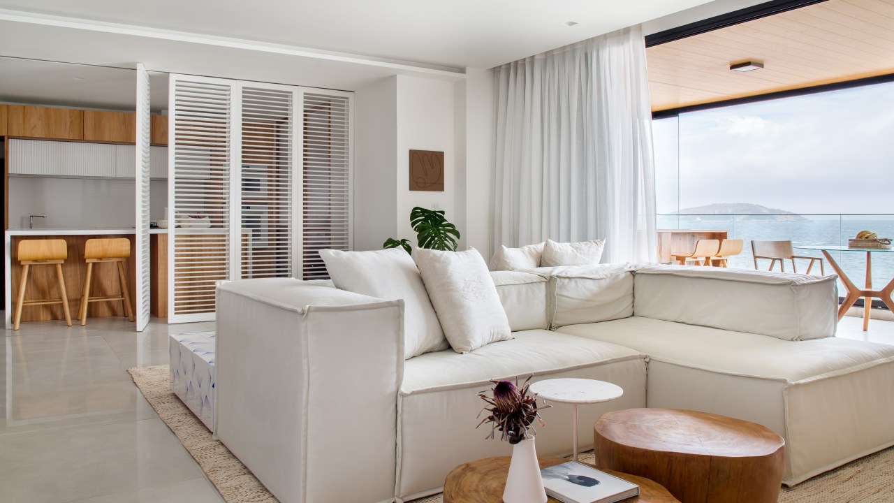 Décor essencial com branco e madeira peroba valoriza o mar neste apê. Projeto de PKB Arquitetura. Na foto, sala de estar, sofá branco, cozinha integrada, veneziana branca.