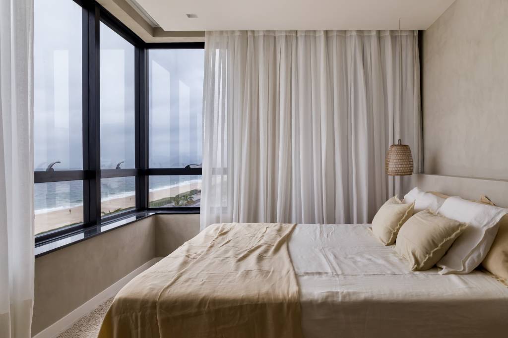 Décor essencial com branco e madeira peroba valoriza o mar neste apê. Projeto de PKB Arquitetura. Na foto, quarto, cortina, roupas de cama bege.