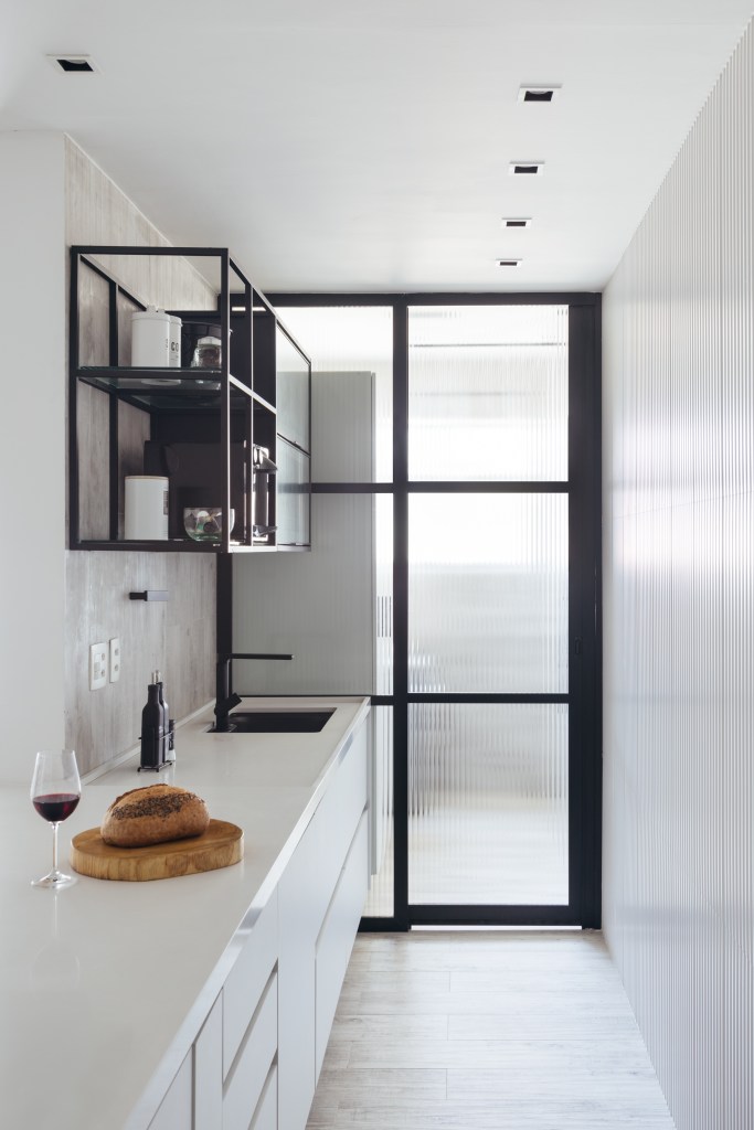 Estilo escandinavo inspira décor de apê de 84m² com paleta preta e branca. Projeto de Nilton Montarroyos. Na foto, cozinha minimalista branca com porta de correr com vidro canelado.