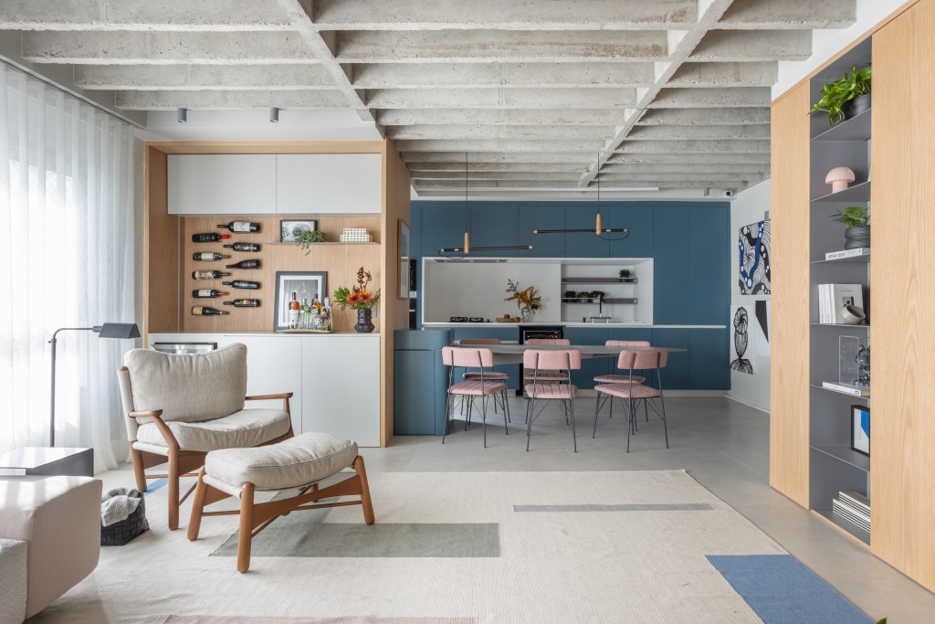 Laje de concreto, mesa em balanço e cozinha azul marcam apê de 115 m². Projeto do escritório Si Saccab. Na foto, cozinha integrada com a sala, adega, marcenaria azul e poltrona.