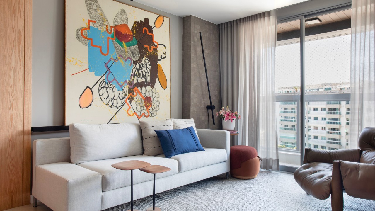Materiais naturais como madeira, couro, linho e fibra natural marcam apê. Projeto de Ikeda Arquitetura. Na foto, sala de estar com sofá cinza claro, quadro colorido acima do sofá.