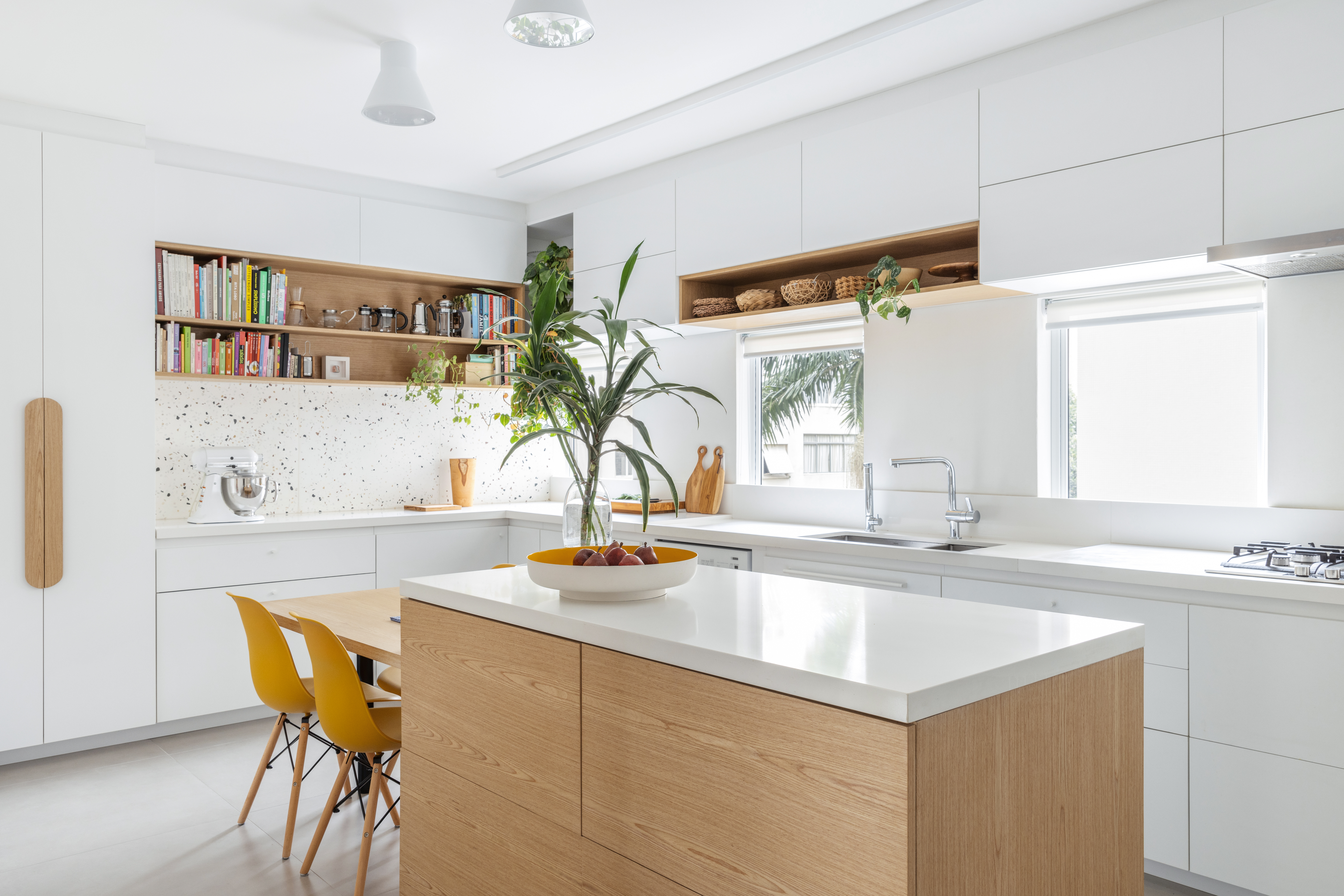 Com paleta branca, apê de 240 m² tem porta de correr inspirada em biombos. Projeto de Lucia Manzano. Na foto, cozinha, bancada branca, marcenaria branca, plantas, cadeiras amarelas.