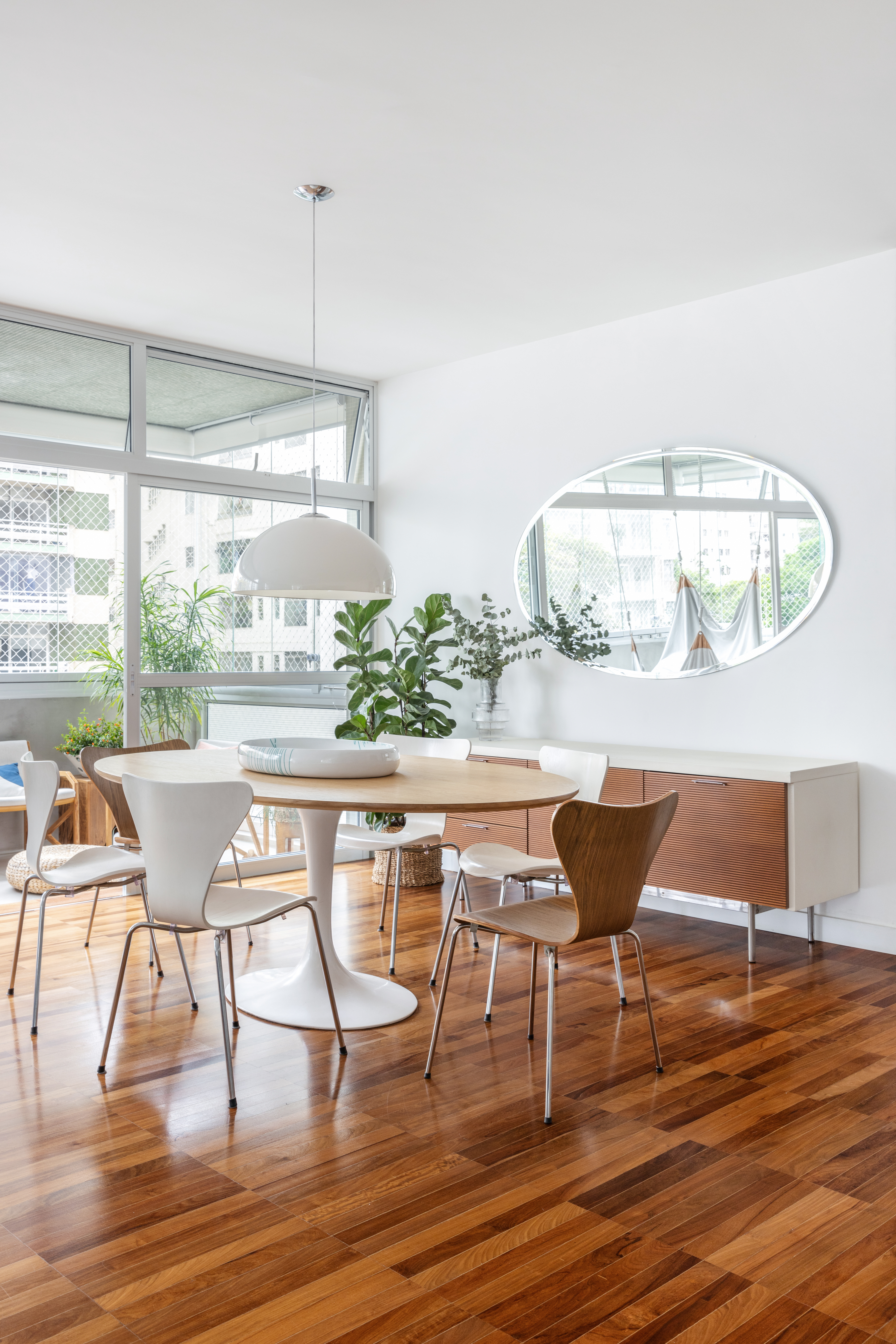 Com paleta branca, apê de 240 m² tem porta de correr inspirada em biombos. Projeto de Lucia Manzano. Na foto, sala, piso de madeira, mesa de jantar, plantas, espelho oval.