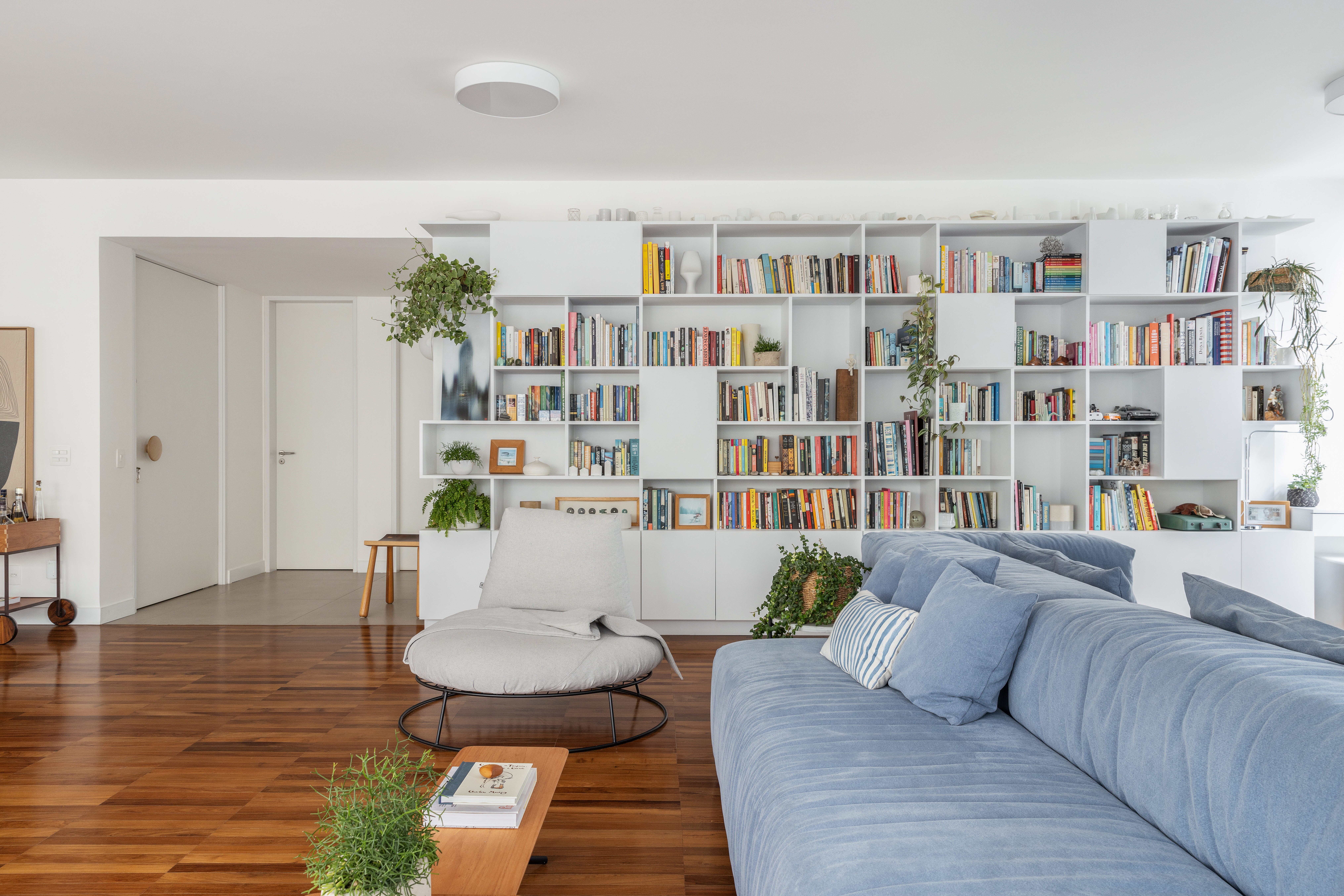 Com paleta branca, apê de 240 m² tem porta de correr inspirada em biombos. Projeto de Lucia Manzano. Na foto, sala, estante, livros, biblioteca, sofá azul.