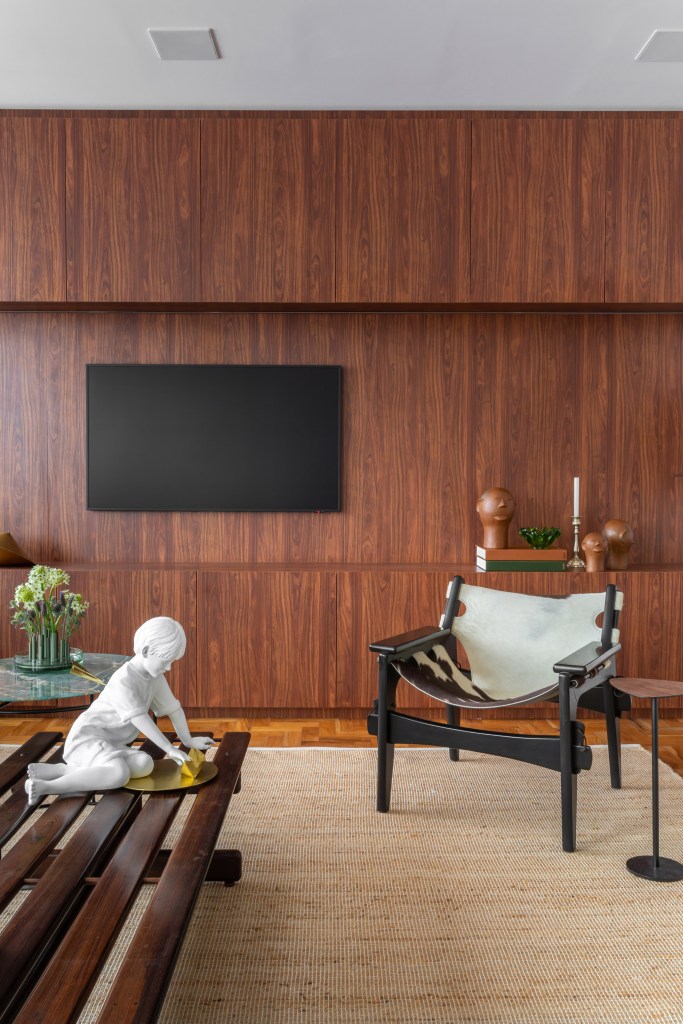 Peças afetivas com memória, design e madeira escura compõem apê de 116 m². Projeto de NOP Arquitetura. Na foto, sala com tv, parede revestida com madeira escura.
