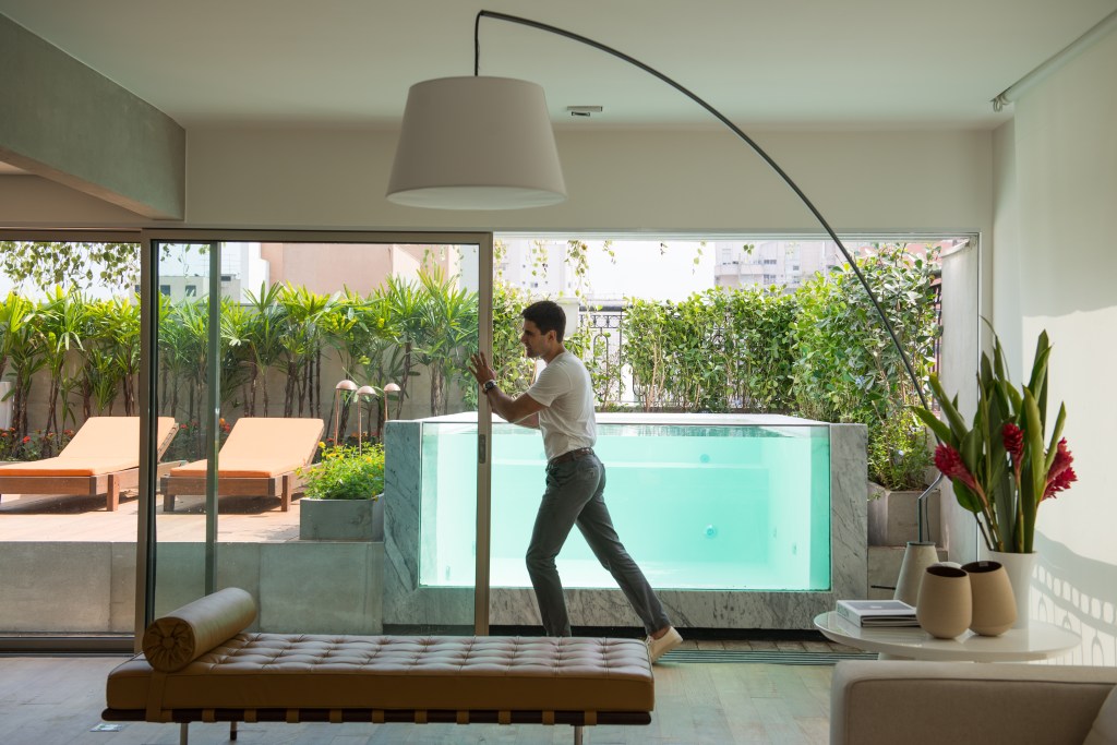 Piscina transparente parece estar dentro da sala neste apê de 400 m². Projeto de Alexandre Del Fabbro. Na foto, portas de vidro separam sala de varanda com piscina.